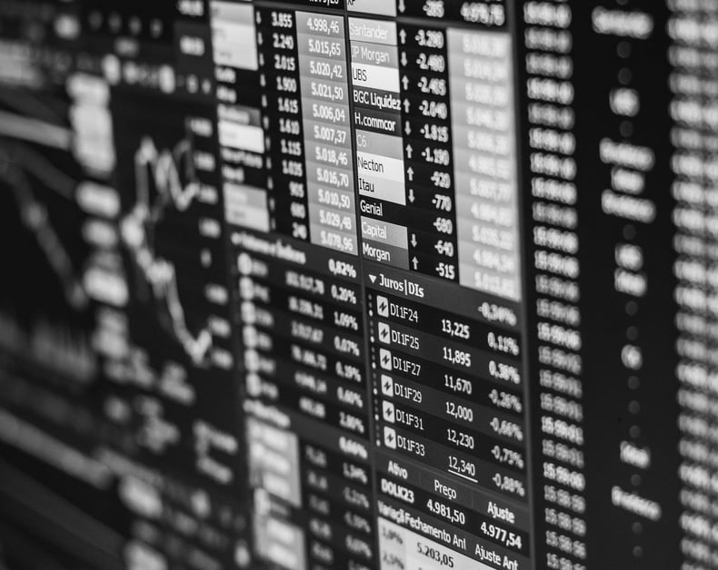 Black-and-white stock market board showing rows of numbers, company names, and tickers.