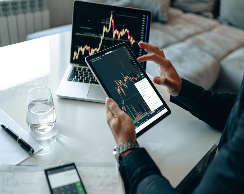 Person using a tablet to view stock charts while a laptop displays a larger chart in the background on a white desk.
