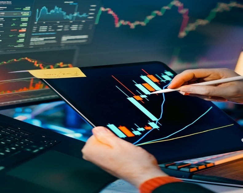 Person uses a tablet with a stock chart and a stylus, laptop nearby, displaying candlestick graphs and a yellow sticky note