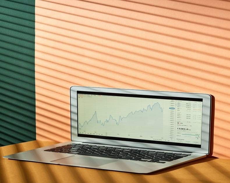 Laptop on a wooden table showing a rising stock chart on the screen with a light grid and data panel on the right side.