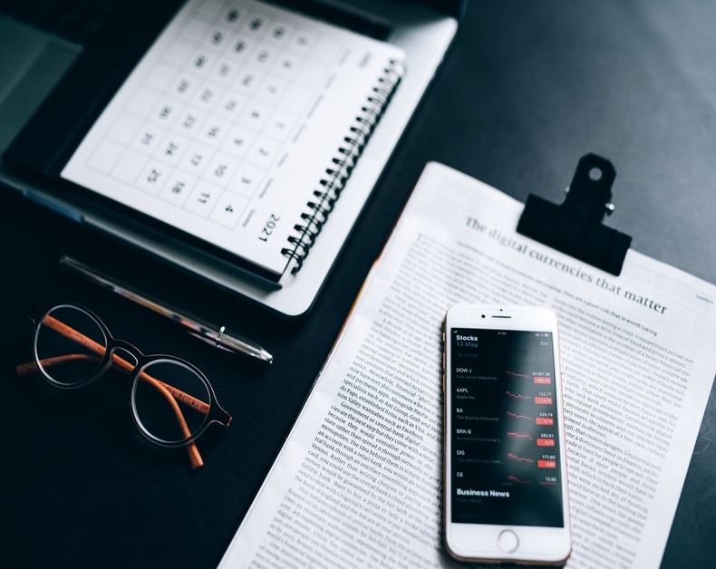 Desk scene with calendar, glasses, pen, and a smartphone showing stock quotes on a newspaper page about digital currencies.