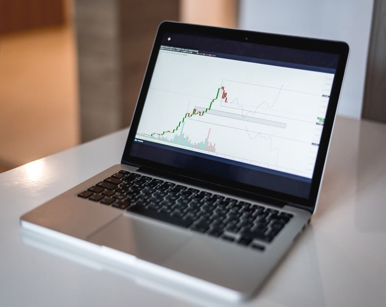 Laptop on a white table showing a stock trading chart with candlesticks and volume bars on the screen