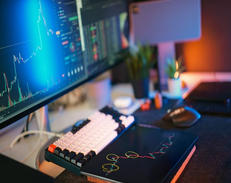 Trading desk with multiple monitors showing stock charts, a white mechanical keyboard, a tablet displaying a candlestick chart, and a mouse on the desk.