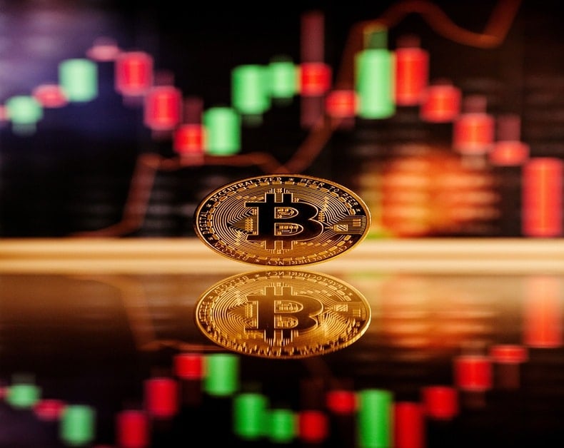 Gold Bitcoin coin spinning on a reflective surface with a colorful candlestick chart in the background as a trading concept image to crypto traders.
