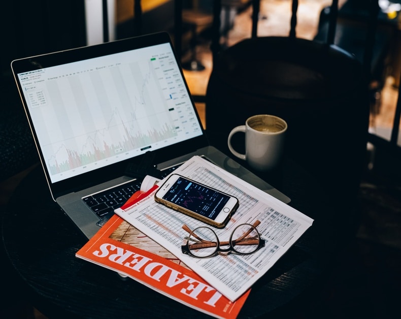 Laptop screen shows a fluctuating stock chart; desk features a coffee mug, glasses, smartphone, and financial papers.